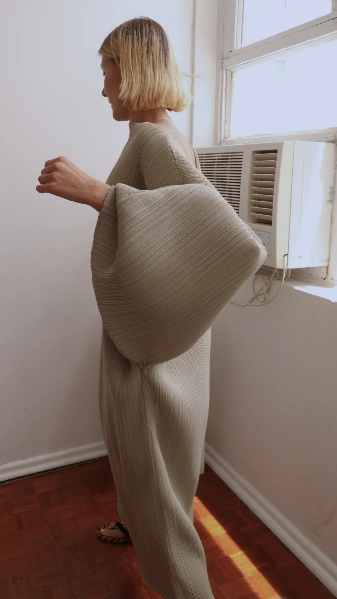 Model beside a window wearing the pleated Patate dress with a sculptural oversized sleeve